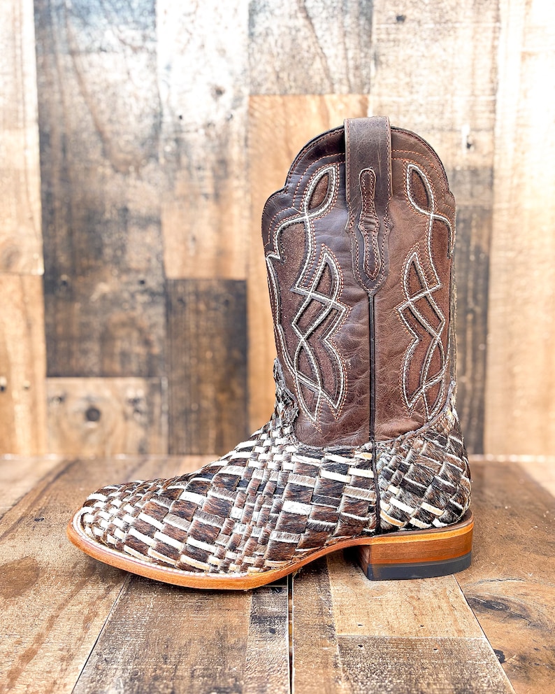 Puede incluir: Una bota de vaquero de cuero marr&oacute;n con un dise&ntilde;o tejido en el pie y una parte superior de cuero marr&oacute;n. La bota tiene costuras decorativas y una puntera cuadrada. El tac&oacute;n mide aproximadamente 3,8 cm de alto.