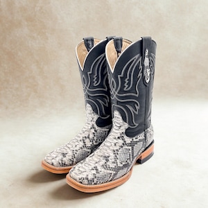 May include: A pair of cowboy boots with a snakeskin pattern on the foot and a black leather upper. The boots have a square toe and decorative stitching. The color scheme is black, white, and brown.