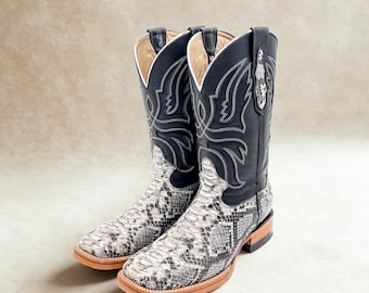 May include: A pair of cowboy boots with a snakeskin pattern on the foot and a black leather upper. The boots have a square toe and decorative stitching. The color scheme is black, white, and brown.