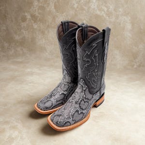 May include: A pair of square-toe cowboy boots featuring a grey and black snakeskin-patterned leather foot and a black leather shaft. The shaft is decorated with intricate white stitching, and the boots have a stacked wooden heel and welt.