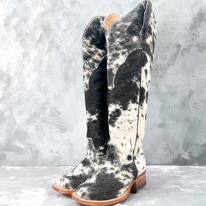 May include: A pair of black and white cowhide knee-high boots with a pointed toe and a low heel.