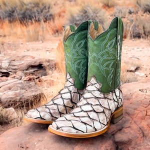 May include: A pair of cowboy boots with a white and brown snakeskin pattern and green leather tops. The boots are sitting on a rock.