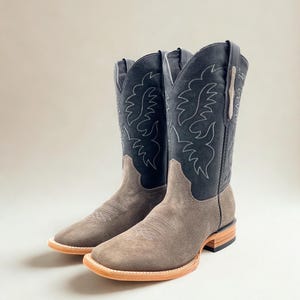 May include: A pair of cowboy boots with a square toe. The boots have a gray suede foot and a black leather upper with white stitching. The soles are light brown.