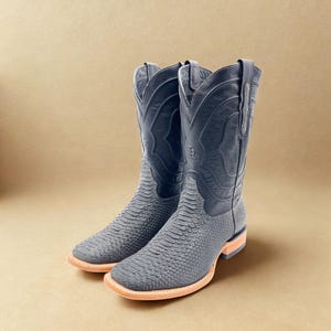 May include: A pair of gray cowboy boots with a textured snakeskin pattern on the foot and a smooth leather upper. The boots have a square toe and a light brown sole. The boots are displayed on a neutral background.
