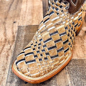 Puede incluir: Primer plano de una bota de estilo western con un dise&ntilde;o tejido. La bota presenta una puntera cuadrada y una parte superior de cuero marr&oacute;n. La secci&oacute;n tejida est&aacute; hecha de materiales beige, negros y blancos.