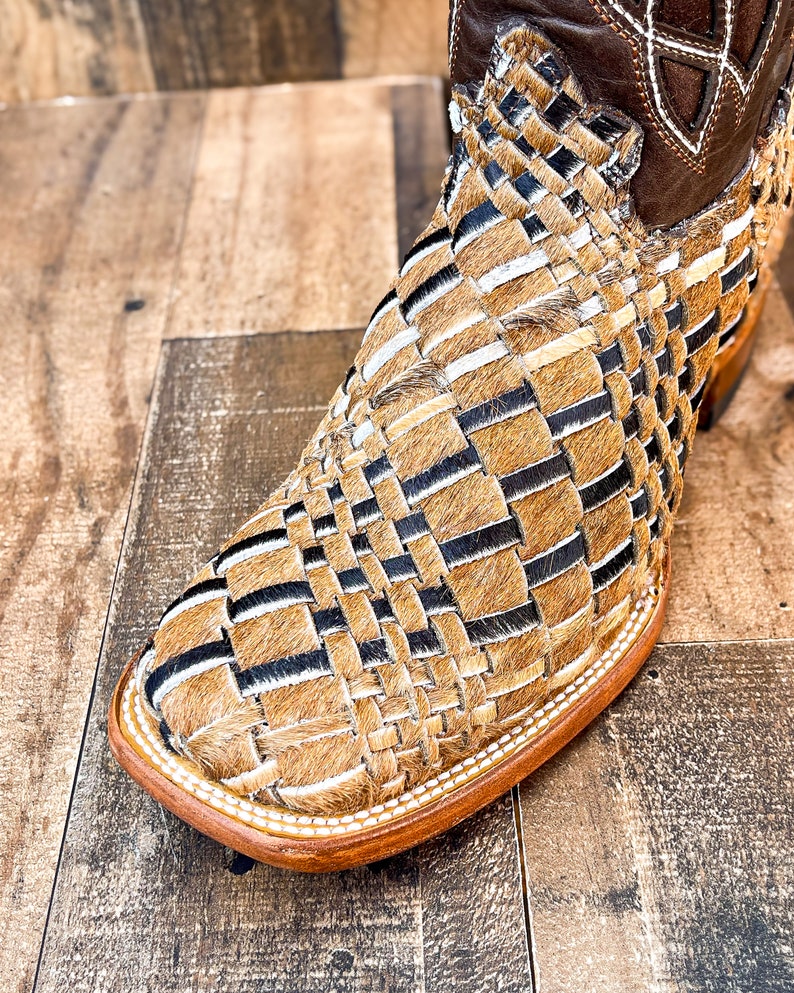 Puede incluir: Primer plano de una bota de estilo western con un dise&ntilde;o tejido. La bota presenta una puntera cuadrada y una parte superior de cuero marr&oacute;n. La secci&oacute;n tejida est&aacute; hecha de materiales beige, negros y blancos.
