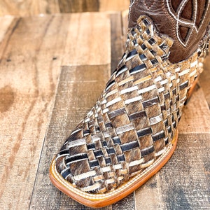 Puede incluir: Primer plano de una bota de vaquero marr&oacute;n y beige. La bota presenta un dise&ntilde;o tejido en el pie y una parte superior de cuero marr&oacute;n con costuras decorativas. La suela es de color marr&oacute;n claro.
