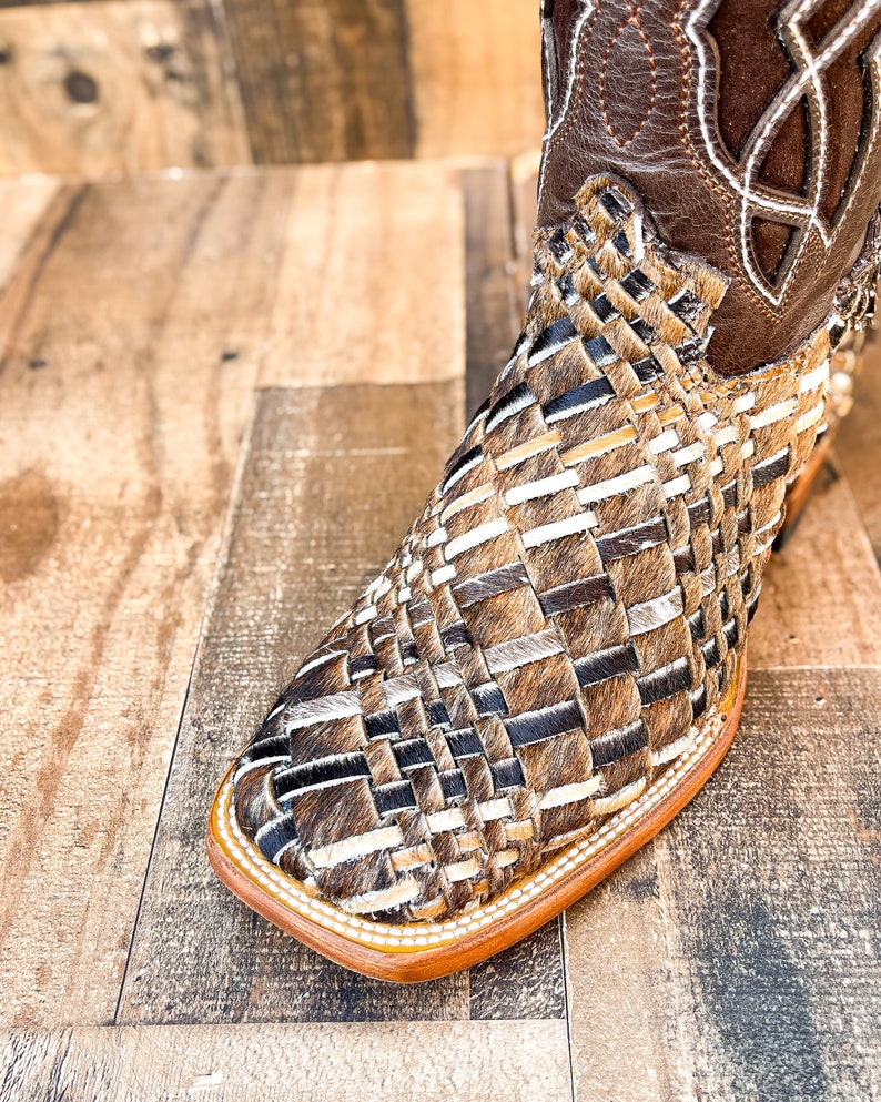 Puede incluir: Primer plano de una bota de vaquero marr&oacute;n y beige. La bota presenta un dise&ntilde;o tejido en el pie y una parte superior de cuero marr&oacute;n con costuras decorativas. La suela es de color marr&oacute;n claro.