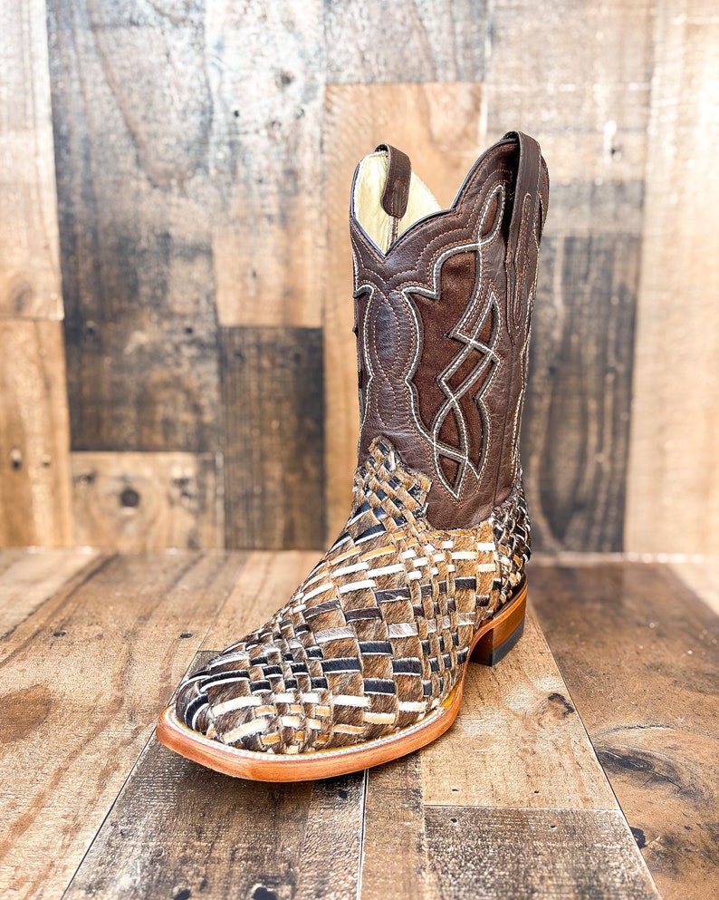 Puede incluir: Una bota de vaquero de cuero marr&oacute;n con un dise&ntilde;o tejido en el pie y un patr&oacute;n cosido decorativo en la ca&ntilde;a. La bota tiene una puntera cuadrada y un tac&oacute;n apilado. La paleta de colores incluye tonos de marr&oacute;n, beige y crema.