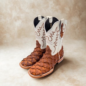 May include: A pair of cowboy boots with a unique design. The boots feature a brown, textured vamp and a white shaft with brown stitching. The boots have a square toe and a stacked heel. The boots are made of leather.