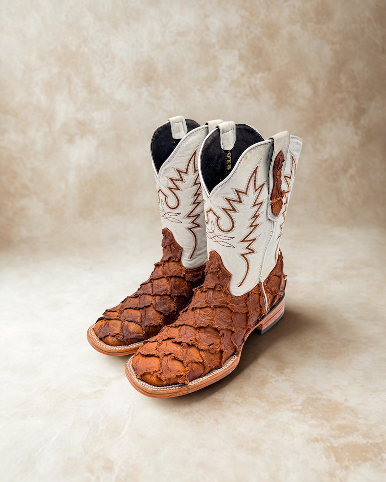 May include: A pair of cowboy boots with a unique design. The boots feature a brown, textured vamp and a white shaft with brown stitching. The boots have a square toe and a stacked heel. The boots are made of leather.