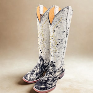 May include: A pair of tall cowboy boots with a cowhide pattern in black and white. The boots have a pointed toe, decorative stitching, and gold-colored studs. The interior is lined with tan leather.
