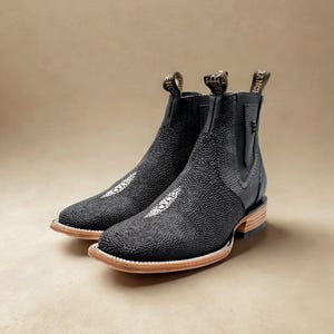 May include: A pair of black leather ankle boots with a textured surface. The boots have a light-colored sole and a pull tab with text. The toe of the boot has a white, speckled design.