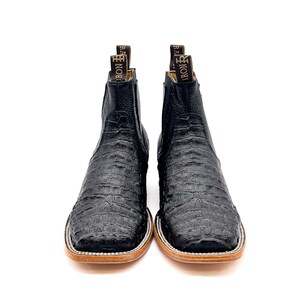May include: A pair of black leather cowboy boots with a textured, crocodile-like pattern. The boots have a square toe, a light brown sole, and a pull tab at the top with the word "IRON" visible. The boots are set against a white background.