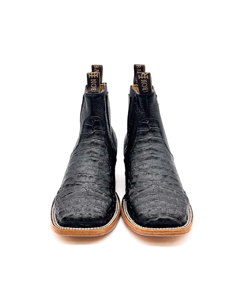 May include: A pair of black leather cowboy boots with a textured, crocodile-like pattern. The boots have a square toe, a light brown sole, and a pull tab at the top with the word "IRON" visible. The boots are set against a white background.