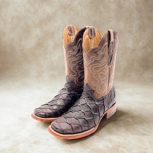 May include: A pair of brown leather cowboy boots. The boots feature a unique scale-like pattern on the foot and a decorative stitched design on the upper shaft. The boots have a square toe and a stacked heel.