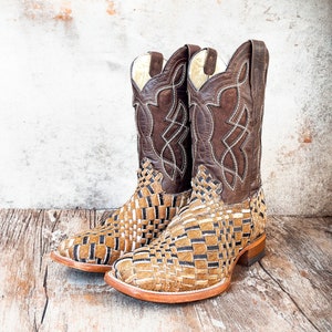 Puede incluir: Un par de botas de vaquero marrones y beige. Las botas tienen un dise&ntilde;o tejido en el pie y una parte superior de cuero marr&oacute;n oscuro con costuras decorativas. Las botas est&aacute;n sobre un fondo de madera r&uacute;stica.