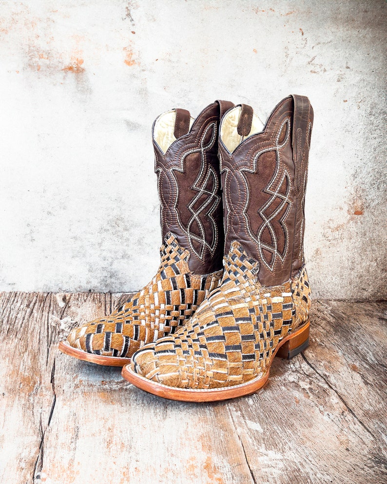 Puede incluir: Un par de botas de vaquero marrones y beige. Las botas tienen un dise&ntilde;o tejido en el pie y una parte superior de cuero marr&oacute;n oscuro con costuras decorativas. Las botas est&aacute;n sobre un fondo de madera r&uacute;stica.