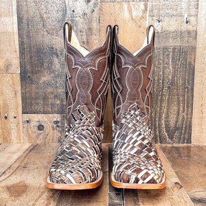 Puede incluir: Un par de botas de vaquero de cuero marr&oacute;n con costuras intrincadas y un dise&ntilde;o tejido en el pie. Las botas tienen una puntera cuadrada y una suela marr&oacute;n claro. La ca&ntilde;a presenta costuras decorativas y un lazo.