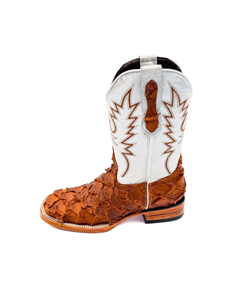 May include: A cowboy boot with a white shaft featuring brown decorative stitching. The vamp is made of textured brown leather. The boot has a square toe and a stacked heel.