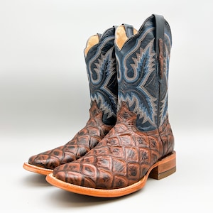 May include: A pair of brown cowboy boots with blue and brown stitching. The boots are made of alligator skin and have a pointed toe.
