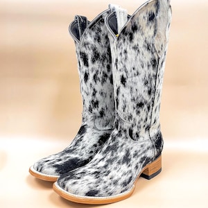 May include: A pair of black and white cowhide cowboy boots with a pointed toe and a low heel.