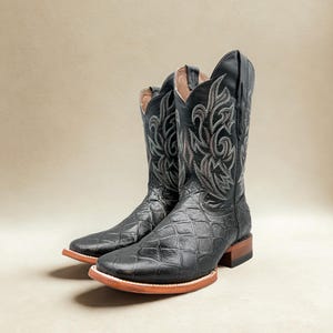 May include: A pair of black cowboy boots with a square toe and a textured pattern. The boots have decorative stitching in shades of gray and brown on the upper shaft. The soles are brown with a light-colored edge.