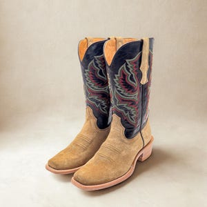 May include: A pair of tan suede cowboy boots with black tops featuring red and green embroidered designs. The boots have a square toe and a stacked heel, showcasing a classic Western style.