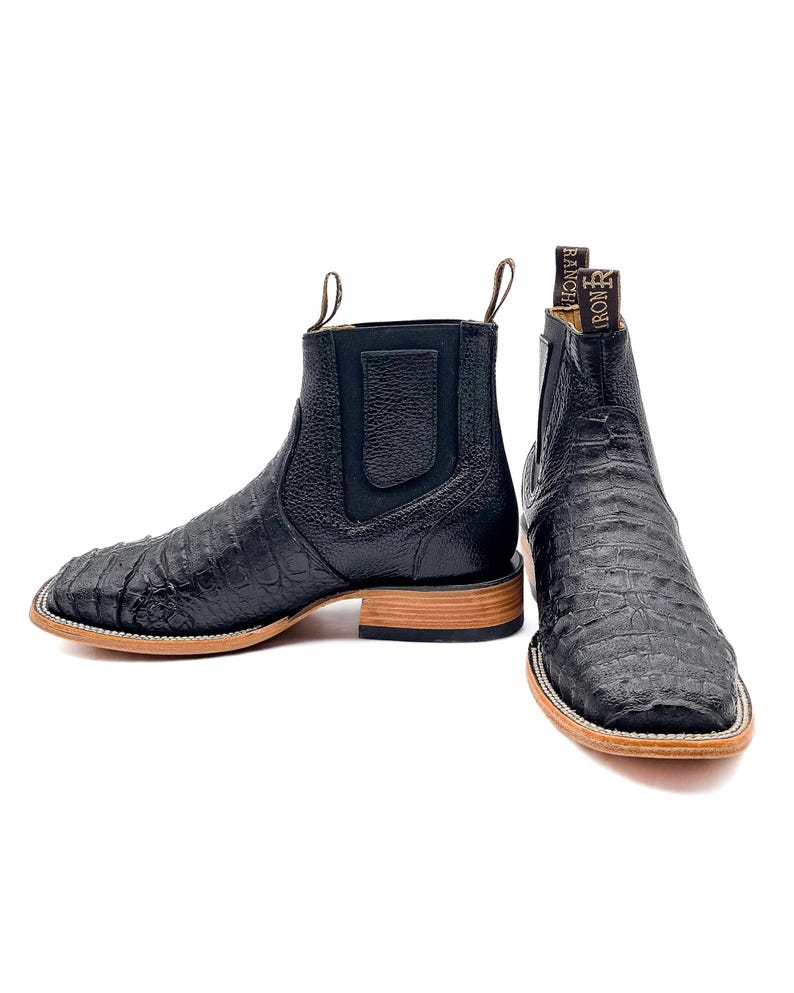 May include: A pair of black leather ankle boots with a square toe and a textured, crocodile-like pattern. The boots have a pull tab at the back and a light brown sole. The brand name "IRON RANCH" is visible on the back of one boot.