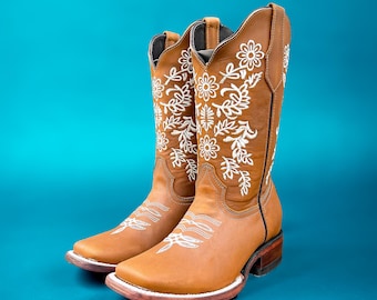 May include: A pair of brown leather cowboy boots with white floral embroidery. The boots have a square toe, a stacked heel, and a tall shaft. The background is a solid teal color.