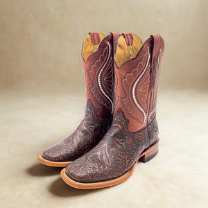 May include: A pair of brown leather cowboy boots with intricate floral and scrollwork designs. The boots have a square toe, a stacked heel, and decorative stitching along the shaft. The interior is a light tan color.