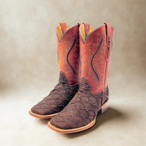 May include: A pair of square-toe cowboy boots featuring a dark brown, textured scale-patterned vamp and a reddish-brown leather shaft. The shaft is decorated with black stitched embroidery and includes pull tabs at the top of each boot.