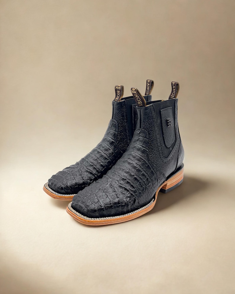 May include: A pair of black leather ankle boots with a textured, crocodile-like pattern. The boots have a square toe, a light brown sole, and a pull tab with the text "IRON RANCH".