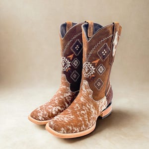 May include: A pair of brown and white cowboy boots. The boots have a square toe and a brown leather upper with decorative stitching and a cowhide pattern on the foot. The boots are approximately 12 inches tall.