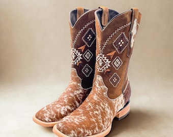 May include: A pair of brown and white cowboy boots. The boots have a square toe and a brown leather upper with decorative stitching and a cowhide pattern on the foot. The boots are approximately 12 inches tall.