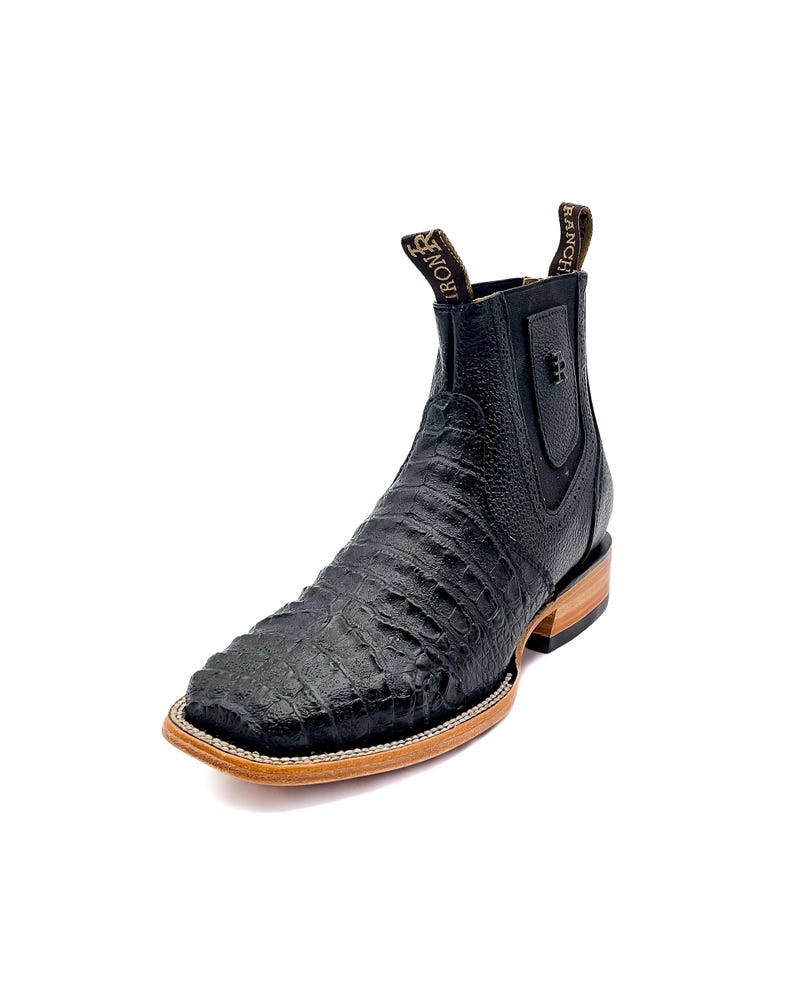 May include: Black leather ankle boot with a square toe and a textured, crocodile-like pattern. The boot has a pull tab with "RANCH IRON" printed on it. The sole is a light brown color with a white stitch.