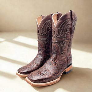 May include: A pair of brown leather cowboy boots with intricate floral and scrollwork designs. The boots have a square toe, stacked heel, and white stitching along the sole. The interior is lined with white leather.