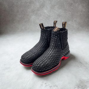 May include: A pair of black ankle boots with a woven design. The boots have a red stripe around the sole and a pull tab at the back. The soles are black and red.