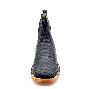 May include: Black leather ankle boot with a square toe and a textured, crocodile-like pattern. The boot has a zipper on the side and a brown sole. The brand name "IRON RIVER" is visible on the pull tab.