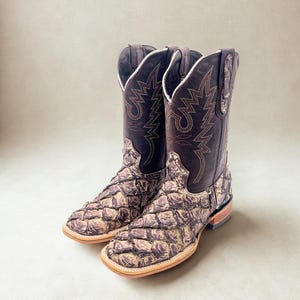 May include: A pair of brown cowboy boots with a unique scale-like texture on the foot and lower leg. The upper shaft is a darker brown with decorative stitching. The boots have a square toe and a light-colored sole.