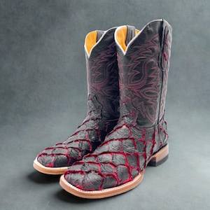 May include: A pair of black cowboy boots with red stitching and accents. The boots have a unique textured pattern on the vamp and shaft, a square toe, and a light brown sole. The interior is lined with a light yellow material.