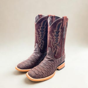 May include: A pair of brown cowboy boots with a square toe. The boots have a textured, scaly pattern on the foot and a suede upper with decorative stitching. The soles are light brown.
