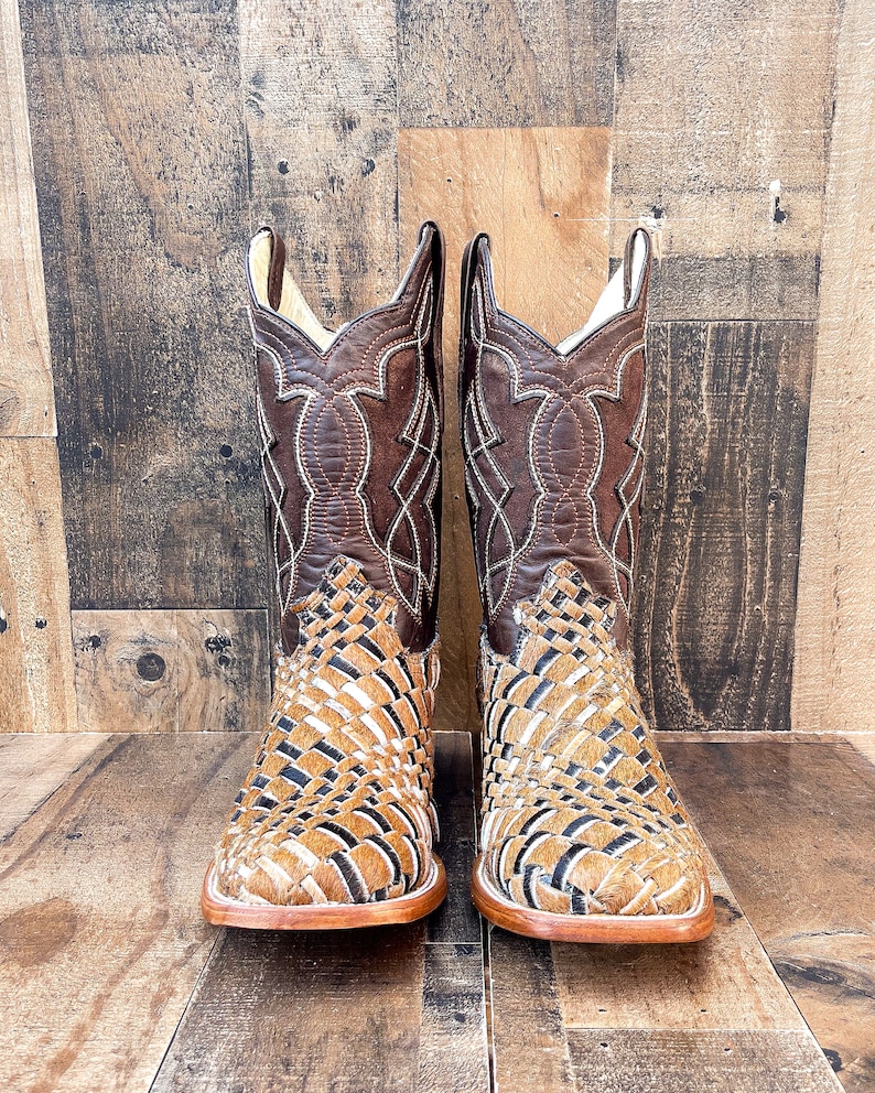 Puede incluir: Un par de botas de vaquero de cuero marr&oacute;n. La parte superior presenta un dise&ntilde;o cosido, mientras que la parte inferior tiene un patr&oacute;n tejido con acentos negros y tostados. Las botas tienen una punta cuadrada y una suela marr&oacute;n claro.