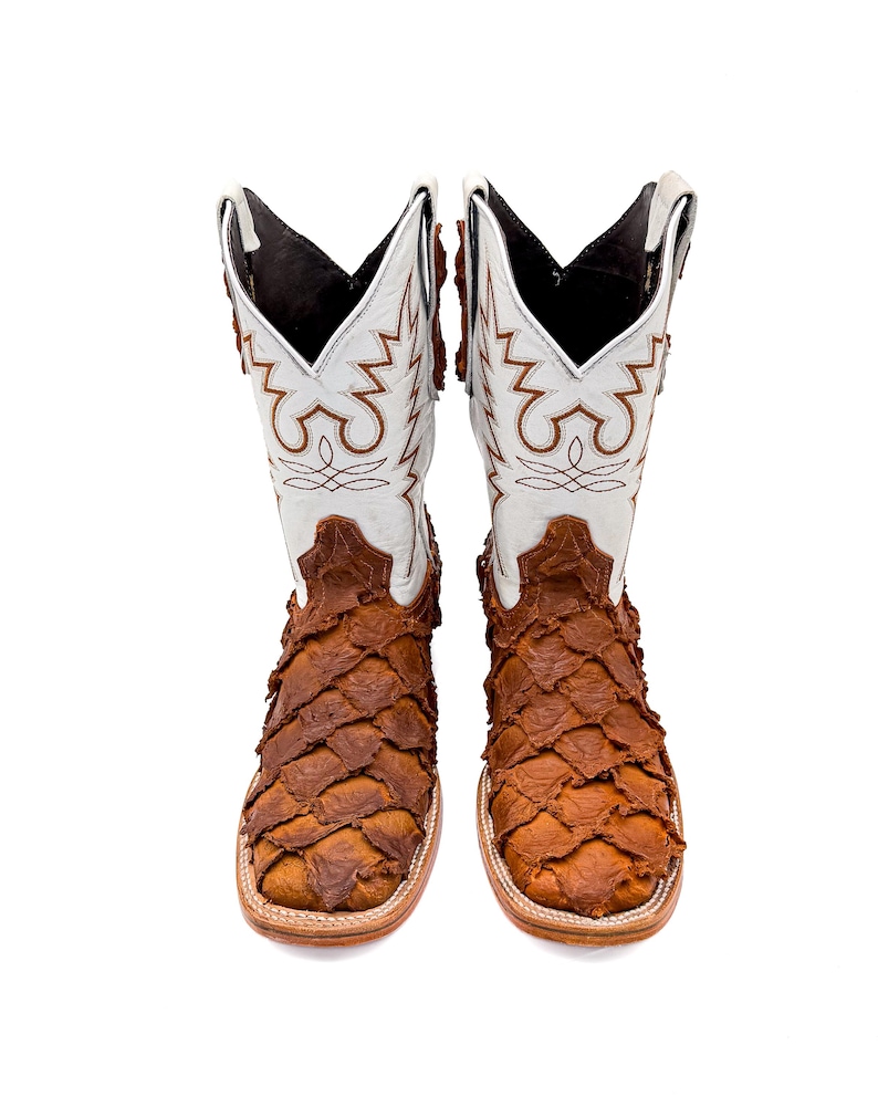 May include: A pair of cowboy boots with a white and brown design. The upper part of the boots is white with brown decorative stitching. The lower part of the boots is brown with a scale-like texture. The boots have a square toe.