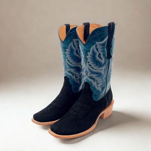 May include: A pair of black suede cowboy boots with blue embroidered shafts. The boots have a square toe, a stacked heel, and a light brown sole. The shaft features a detailed floral design.