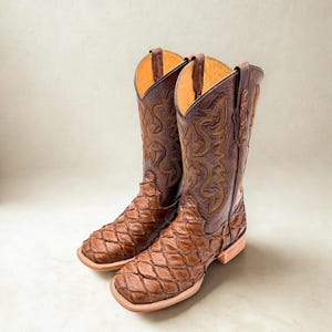 May include: A pair of brown cowboy boots featuring a textured, scale-like pattern on the foot and a smooth leather shaft with decorative gold-toned stitching. The boots have a square toe, a stacked heel, and pull tabs at the top of the shaft.