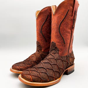 May include: A pair of brown cowboy boots with a unique textured design on the foot and a smooth, reddish-brown leather upper. The boots feature decorative stitching and a square toe, perfect for a Western-inspired look.