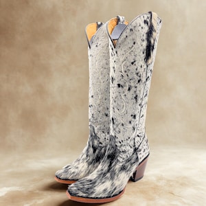 May include: A pair of tall cowboy boots with a spotted black and white pattern. The boots have a pointed toe, a brown heel, and decorative stitching on the upper shaft. The boots are made of a textured material.