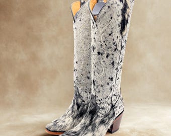 May include: A pair of tall cowboy boots with a spotted black and white pattern. The boots have a pointed toe, a brown heel, and decorative stitching on the upper shaft. The boots are made of a textured material.