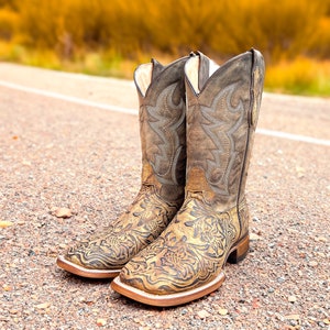 May include: A pair of brown leather cowboy boots with intricate floral embroidery. The boots have a pointed toe and a low heel.
